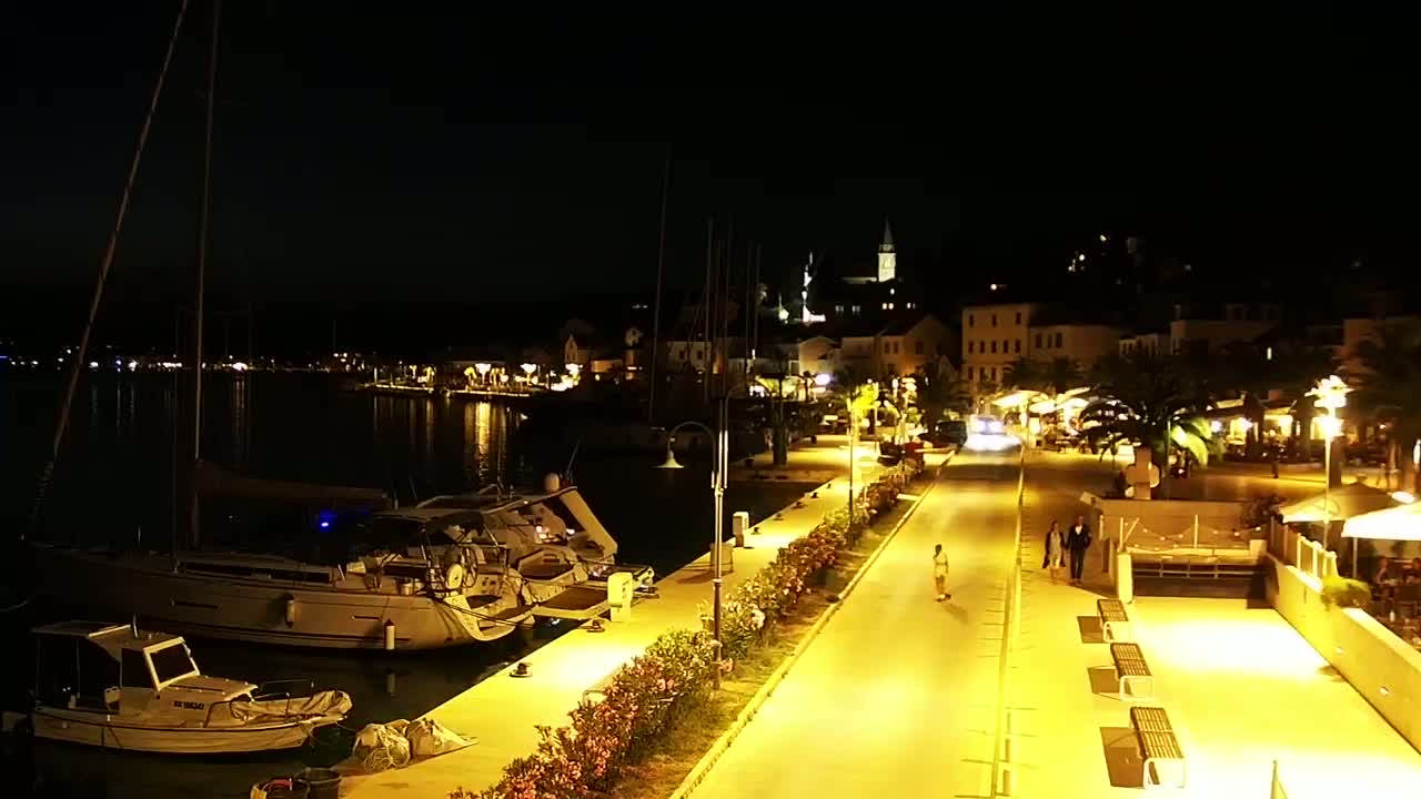 A nighttime view of a harbor town's waterfront, showing boats docked along a brightly lit walkway leading to buildings, under a clear, dark sky.