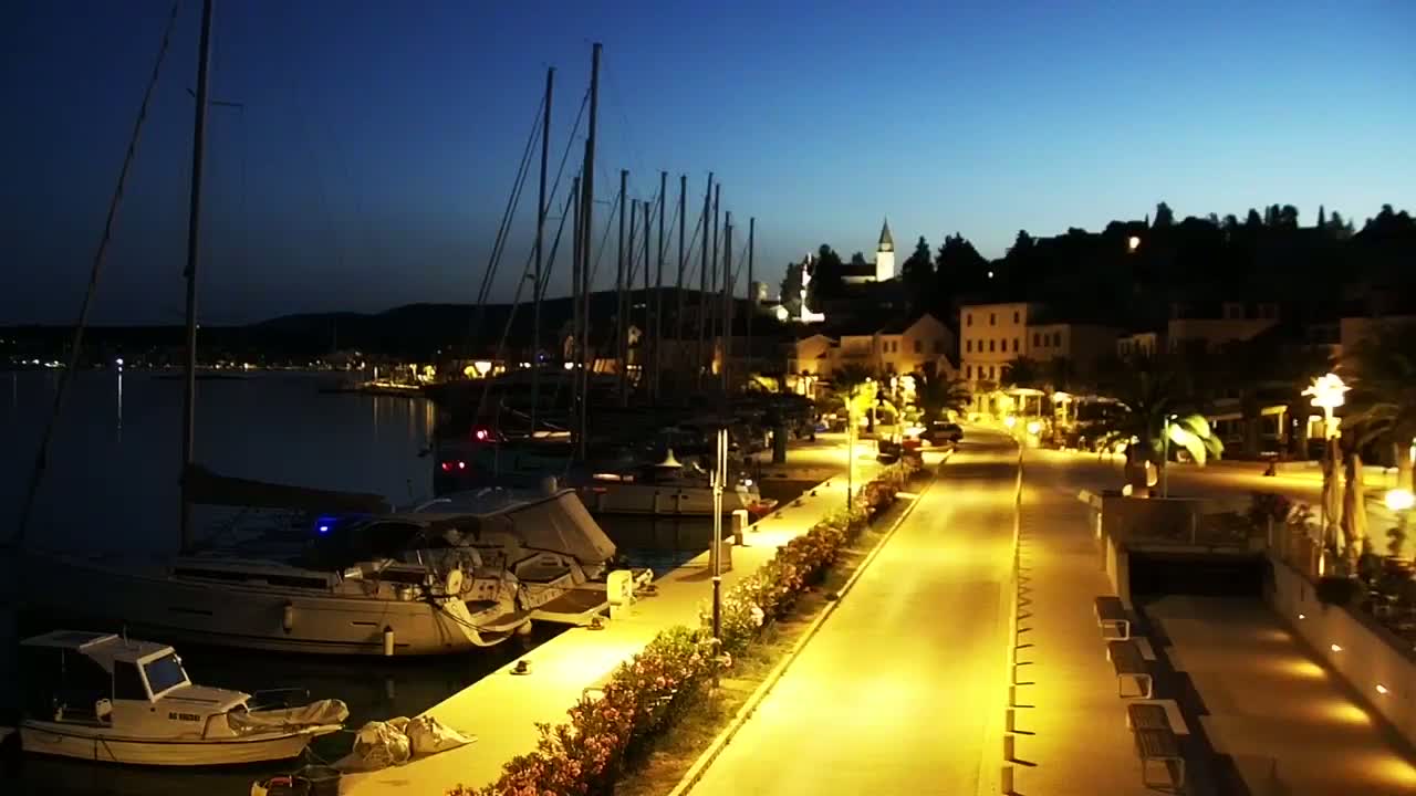 A brightly lit waterfront promenade at night, lined with docked sailboats and overlooking a small, illuminated town nestled against a hill under a dark blue sky.