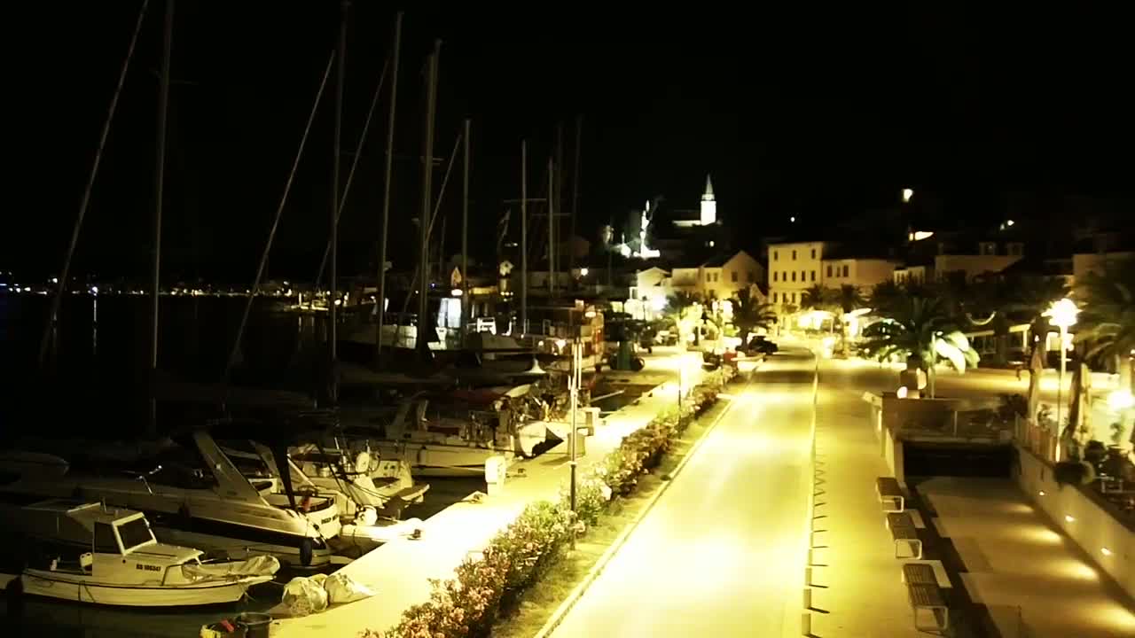 A nighttime view of a harbor with boats docked along a brightly lit waterfront promenade leading to a village.