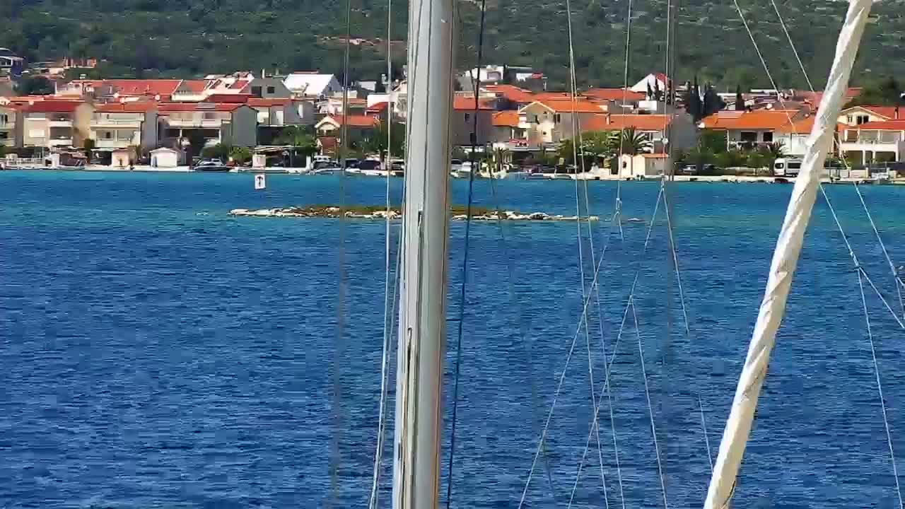 A small island is visible in the calm blue water, with a coastal town of red-roofed houses in the background on a sunny day.