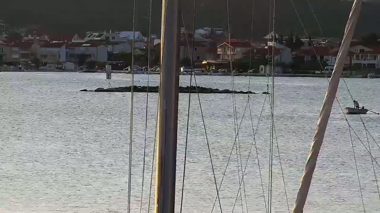 A calm body of water with a small rowboat and person, viewed from behind the masts of a sailboat, shows a shoreline with buildings in the background.