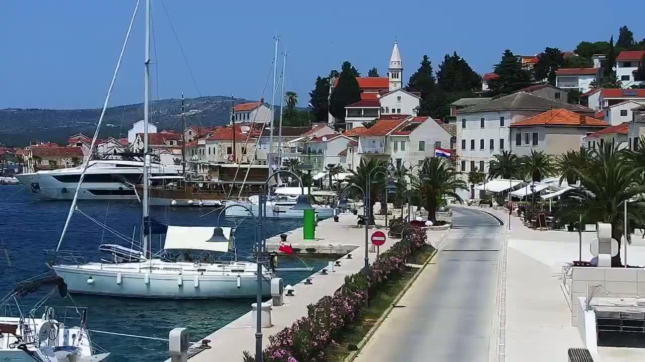 A waterfront town with red-roofed buildings, palm trees, and several sailboats and yachts in a harbor on a sunny day.