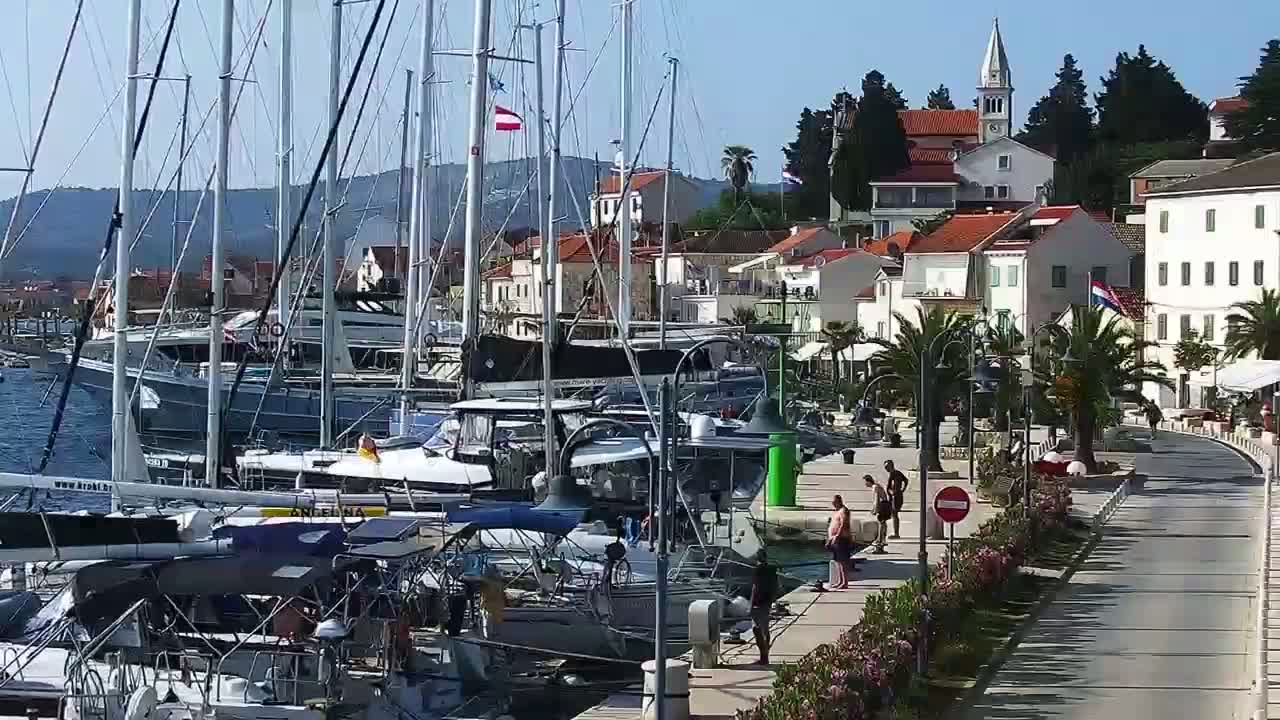 A marina filled with sailboats and motorboats is situated next to a waterfront town under a sunny sky.