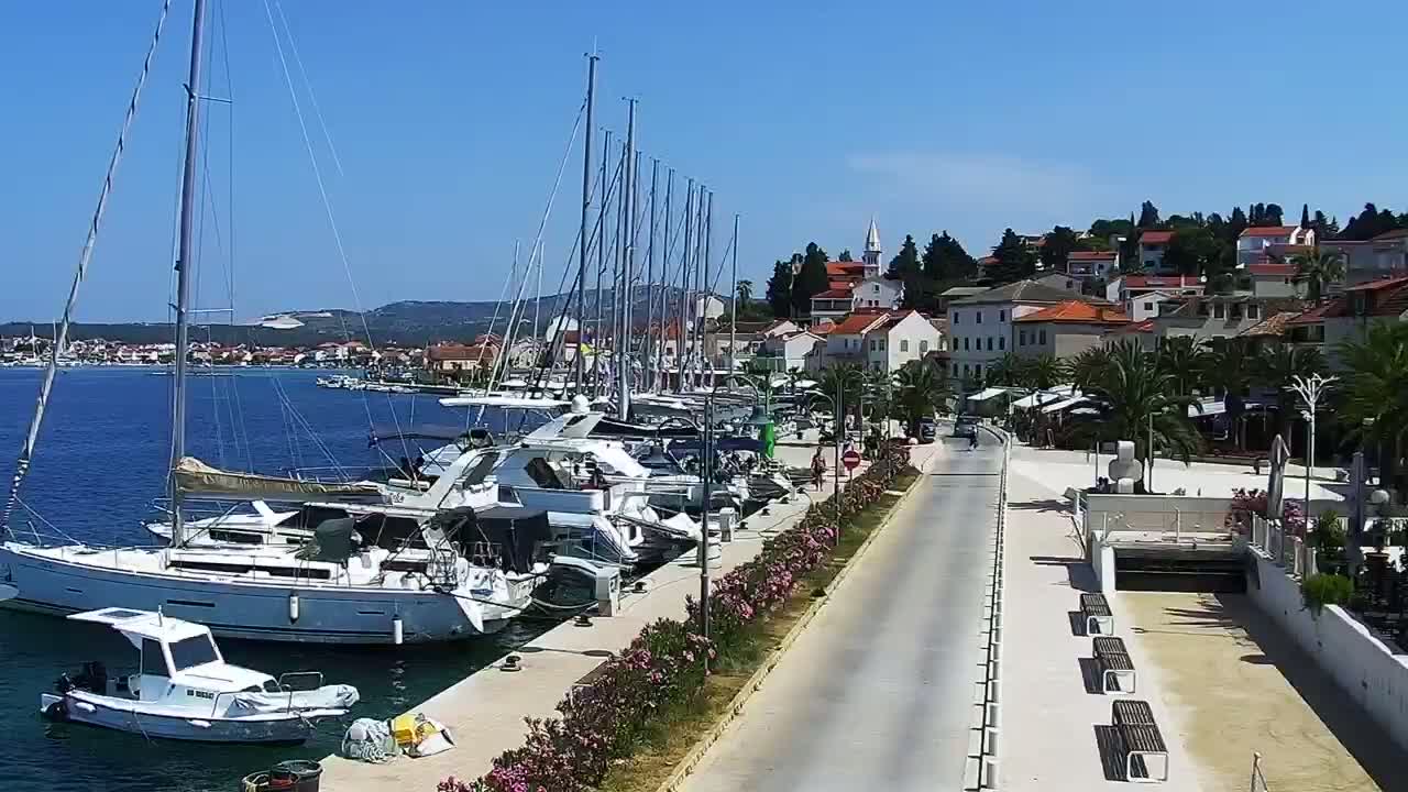 A waterfront town on a sunny day, with numerous sailboats and motorboats docked along a paved walkway that leads to buildings and palm trees.