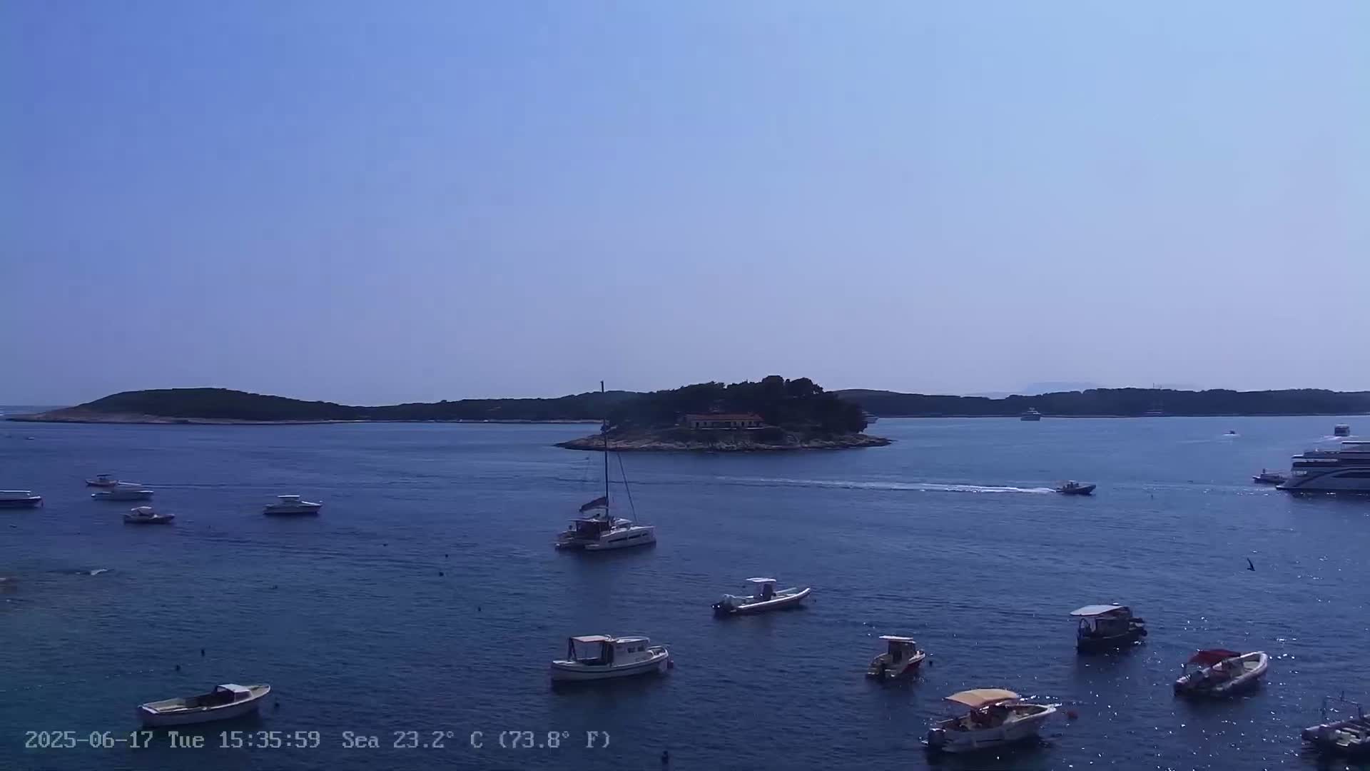 Numerous small boats are scattered across a calm, blue sea in front of a small island with a building on it, under a clear, sunny sky.