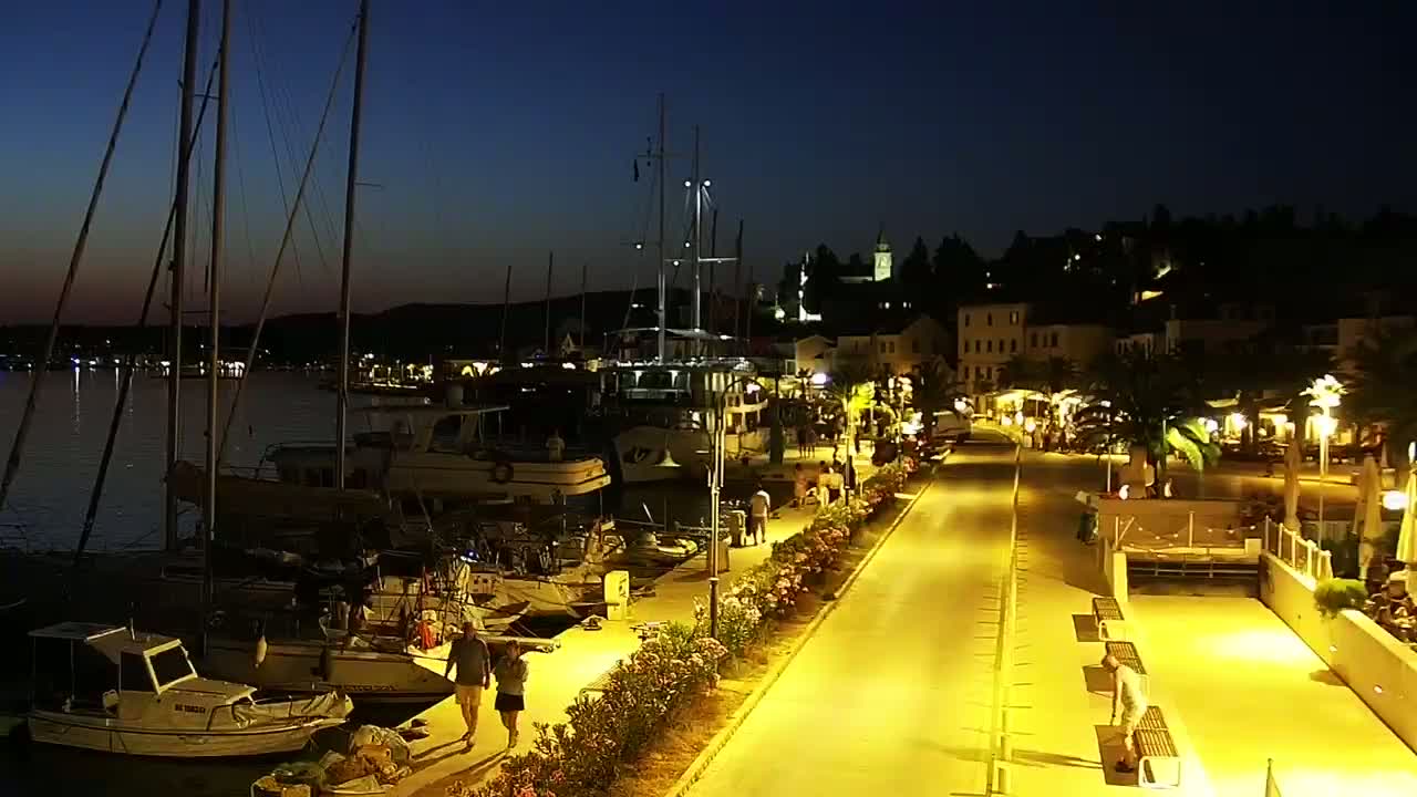 A nighttime harbor scene shows sailboats moored along a paved waterfront walkway lined with lights and flowering plants, under a dark blue sky.
