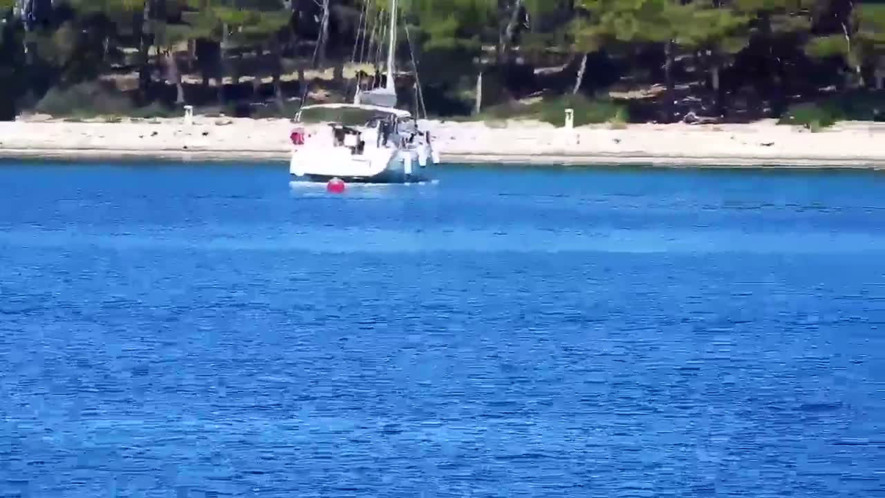 A sailboat with several people aboard is anchored in calm blue water near a sandy shore lined with green trees.