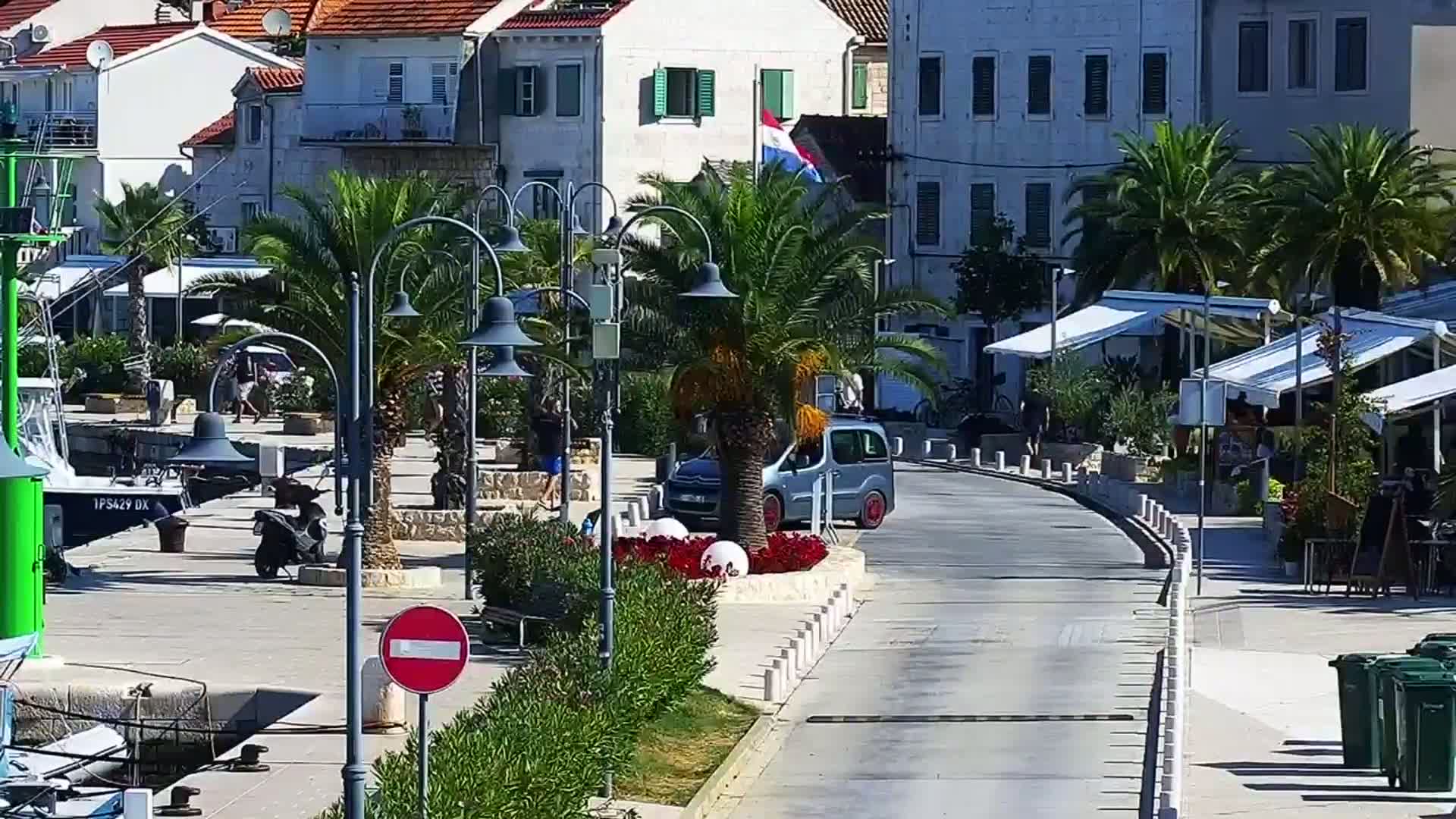A sunny waterfront scene shows a paved road curving past buildings with palm trees, outdoor seating, and a small car parked near a harbor with boats.