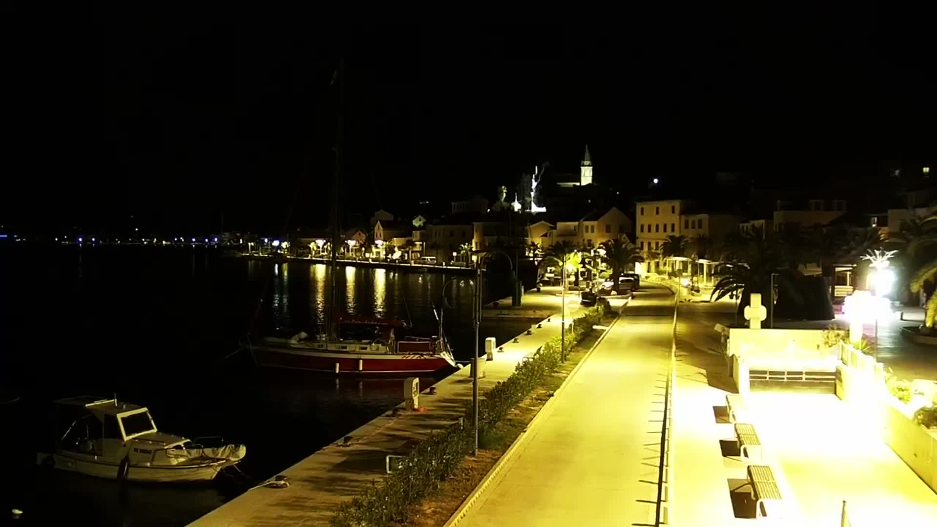 A clear night reveals a brightly illuminated waterfront village with boats moored in the calm dark water, a vibrant promenade, and buildings leading up to a church tower on the hill.