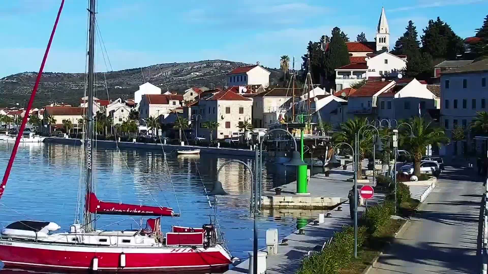 A red sailboat is docked in calm blue waters next to a sunny promenade lined with palm trees and a picturesque coastal town featuring white and terracotta-roofed buildings, a prominent church spire, and distant hills, all under a clear blue sky.
