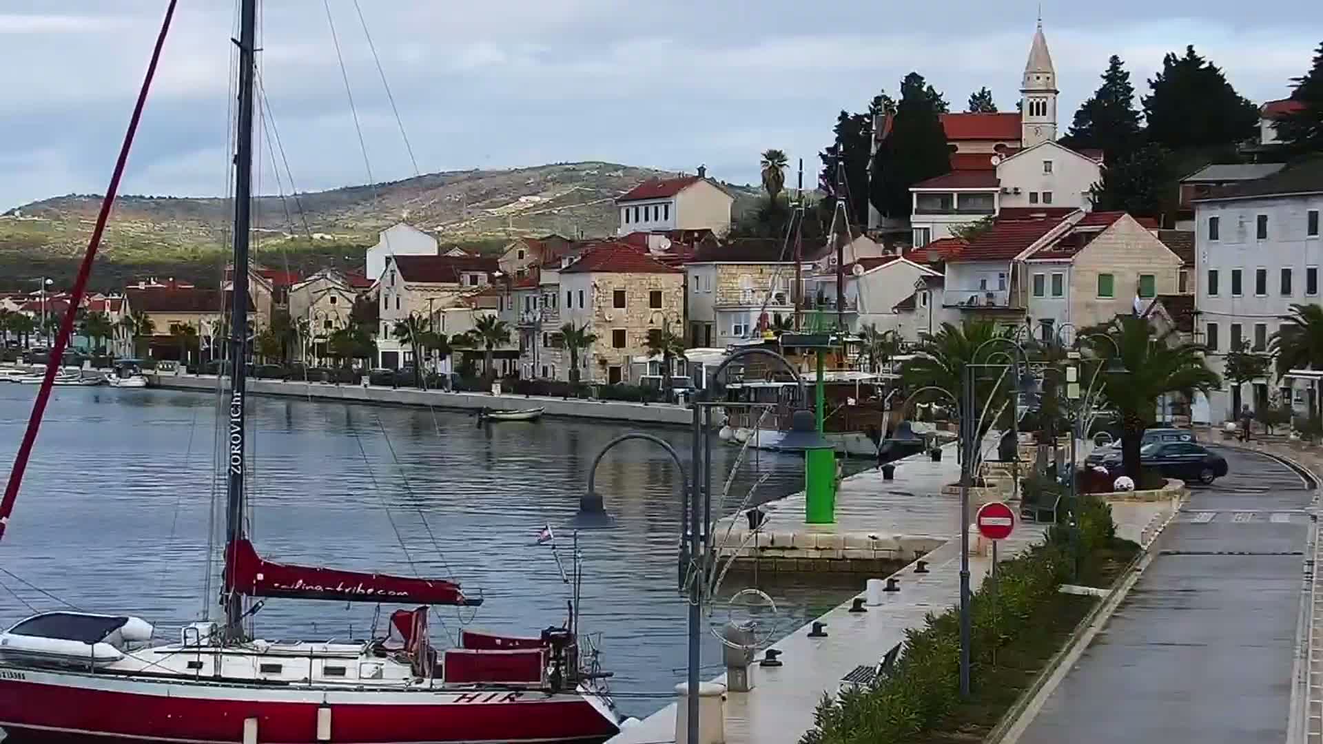 A red and white sailboat is docked in a calm harbor on an overcast day, facing a coastal town with traditional buildings and a prominent church tower, all set against a backdrop of distant hills.