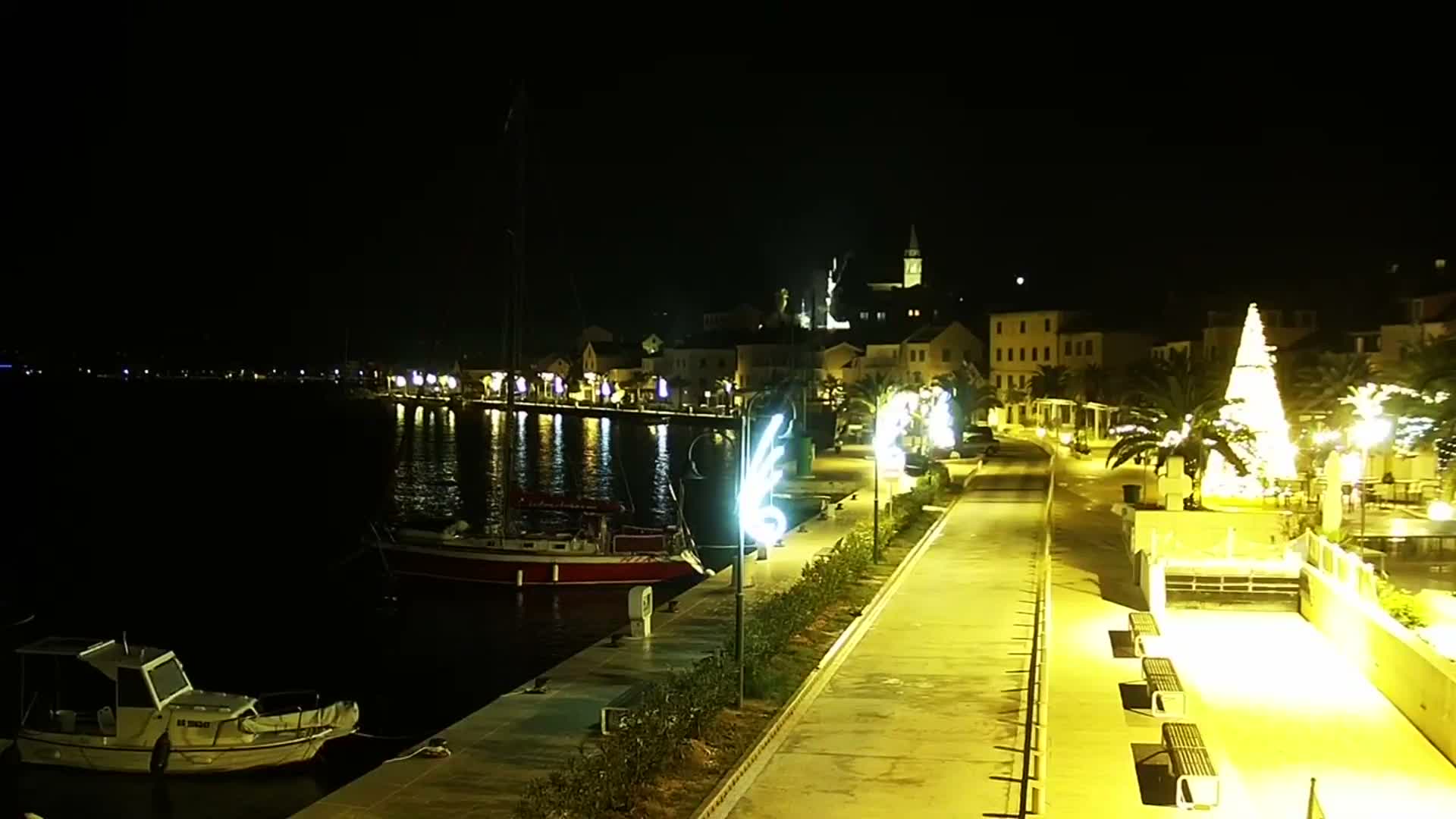 Under a clear night sky, a festive waterfront town glows with illuminated buildings, palm trees, and a large Christmas tree along a promenade, all reflecting on the dark water where boats are docked.
