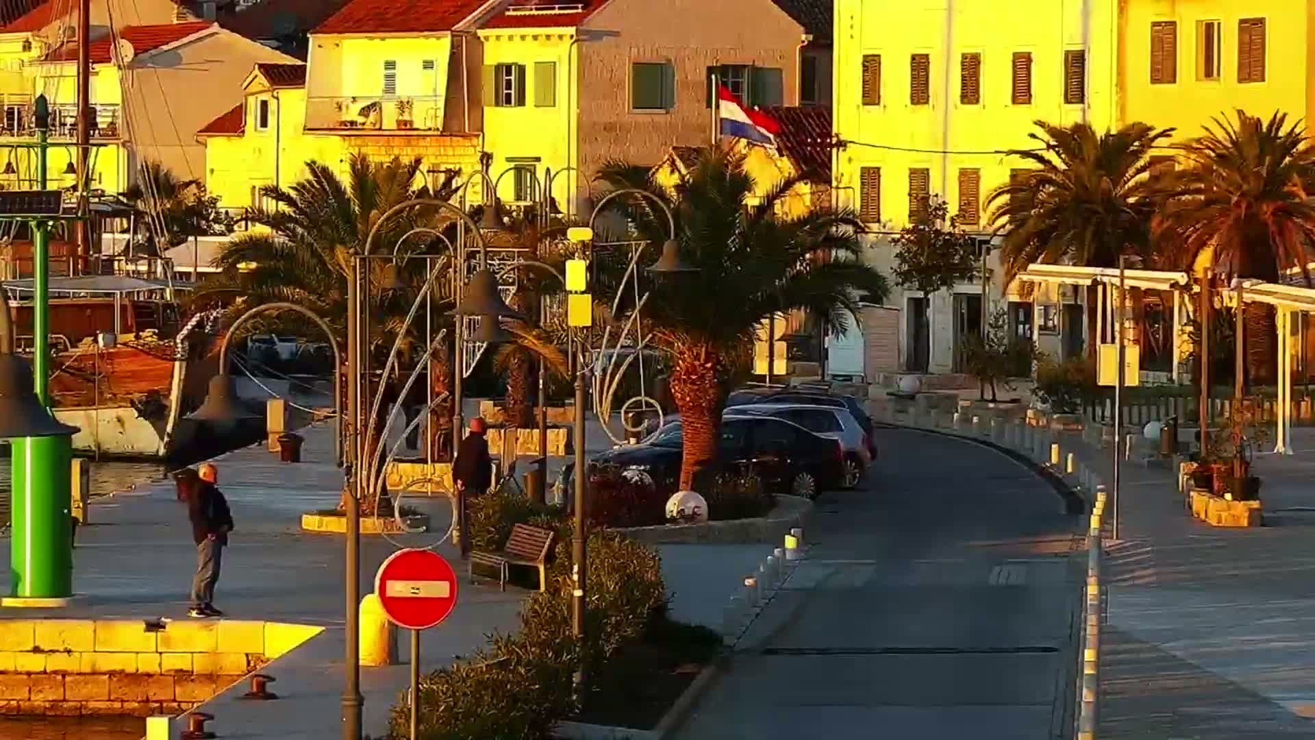 Under a clear night sky, a festive waterfront town glows with illuminated buildings, palm trees, and a large Christmas tree along a promenade, all reflecting on the dark water where boats are docked.