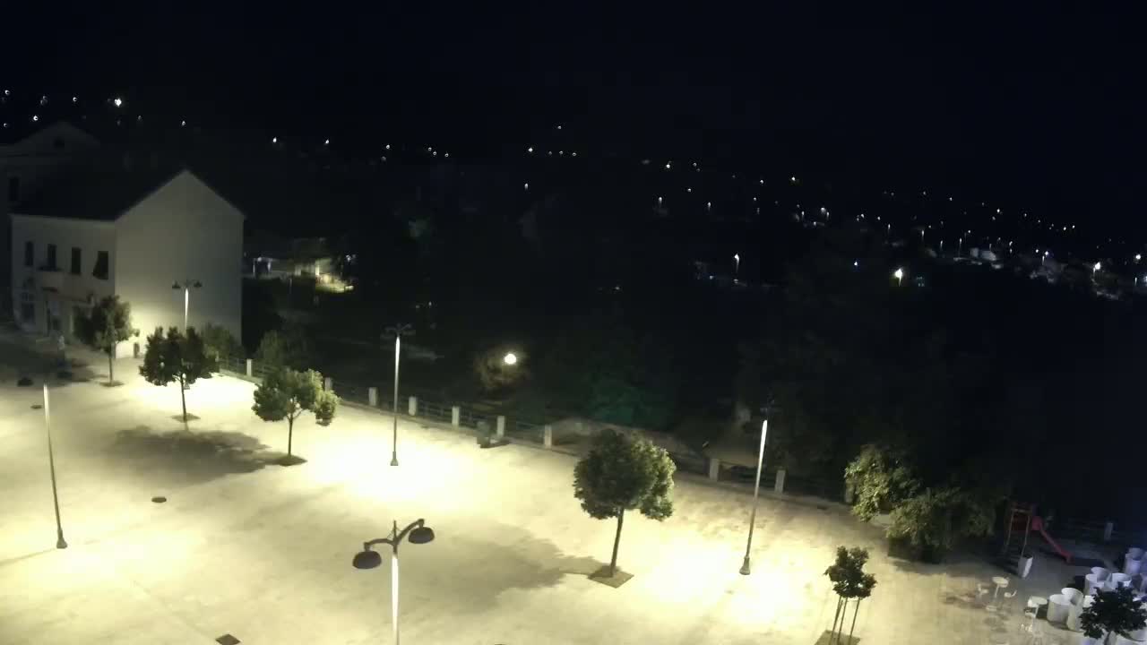 An empty, brightly lit plaza at night, surrounded by trees and buildings, overlooks a dark, sparsely lit residential area.