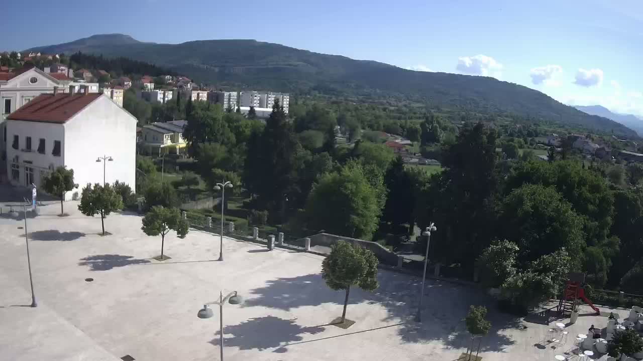A mostly sunny view of a town square with several trees, buildings, and a park, situated at the base of a mountain range.