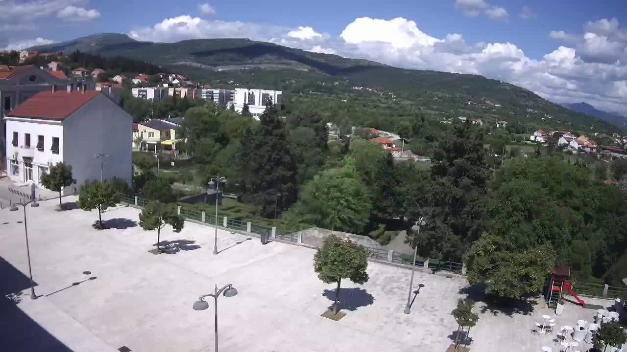 A mostly sunny day reveals a town square with several trees and lampposts, overlooking a verdant valley and mountains in the distance.