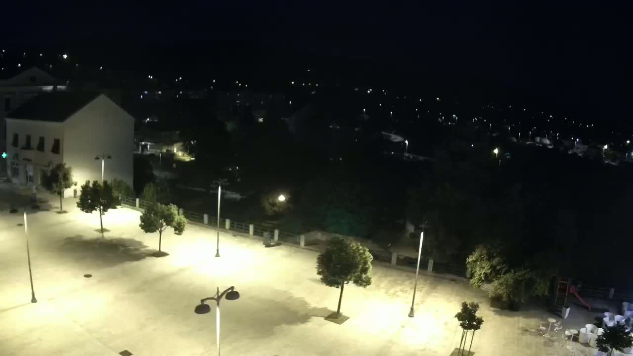 An empty town square at night, illuminated by lampposts, is surrounded by buildings and trees, with a small playground and outdoor seating area visible in the lower right.