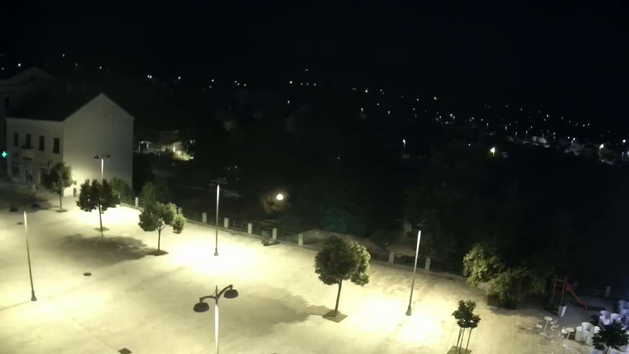 An empty, brightly lit plaza at night, surrounded by buildings and trees, overlooks a dark, sparsely lit town.