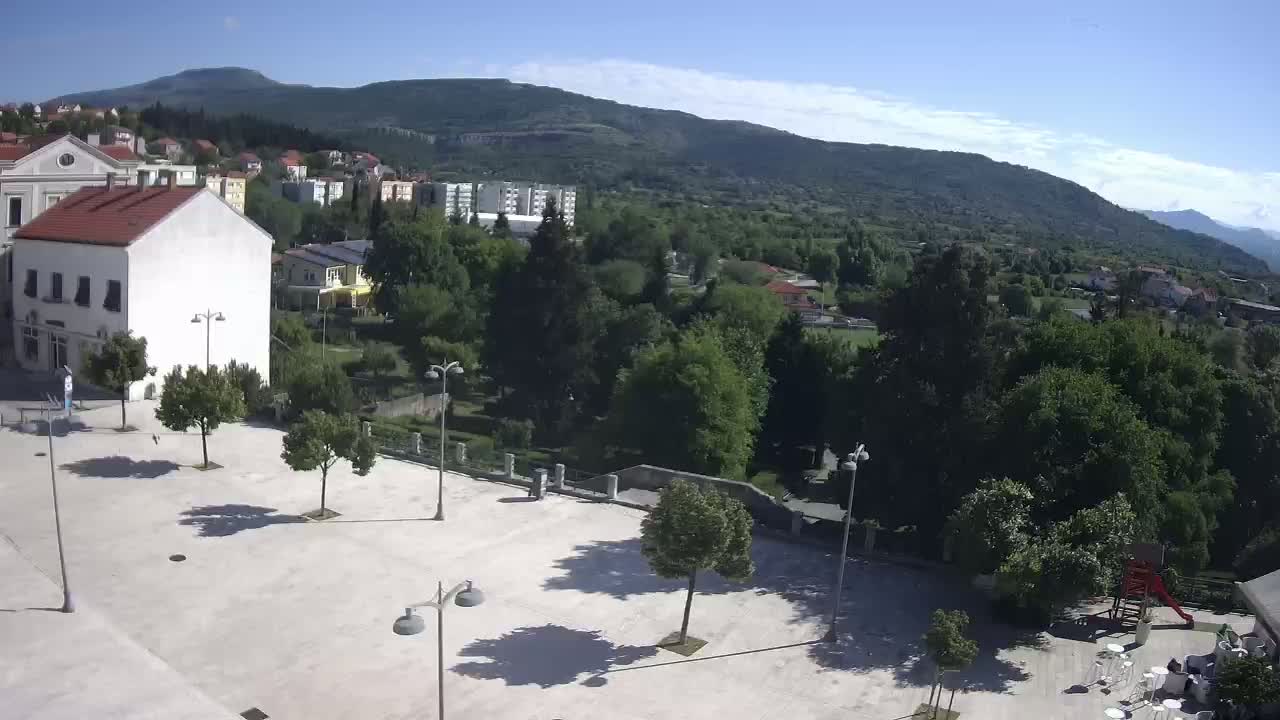 A sunny day reveals a town square with several trees and lampposts, overlooking a verdant hillside and distant mountains.