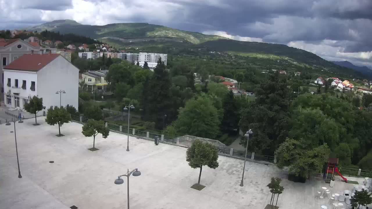 A mostly empty paved square with a few trees and streetlights is situated in front of buildings and a green, hilly landscape under a partly cloudy sky.