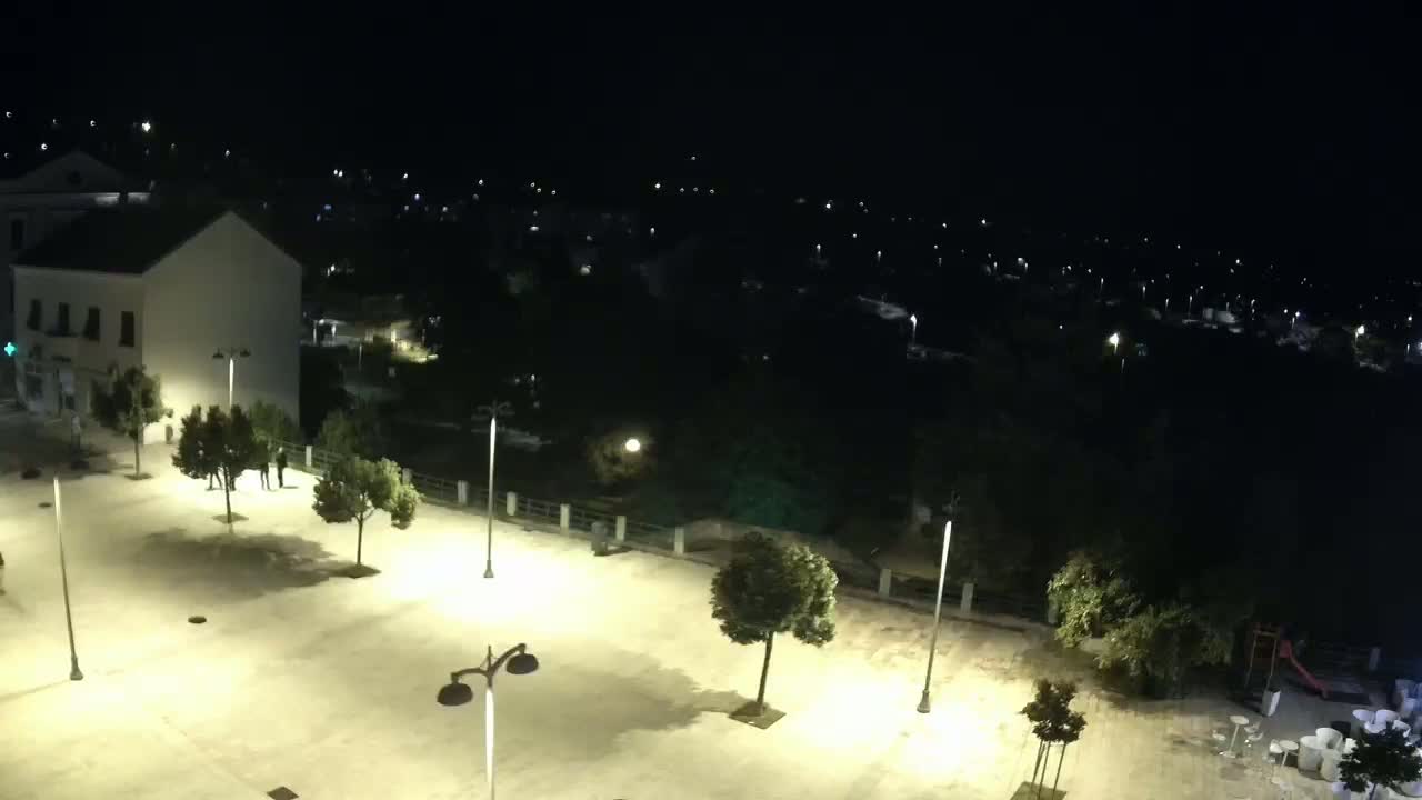 A brightly lit town square at night, surrounded by trees and buildings, overlooks a darker, more densely populated area beyond.