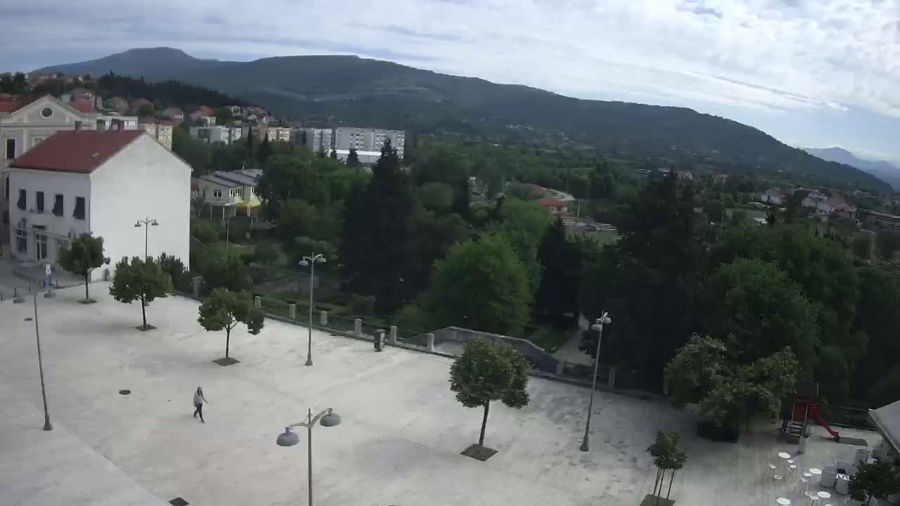 An empty town square with a few people and trees, overlooking a hilly landscape under a cloudy sky.