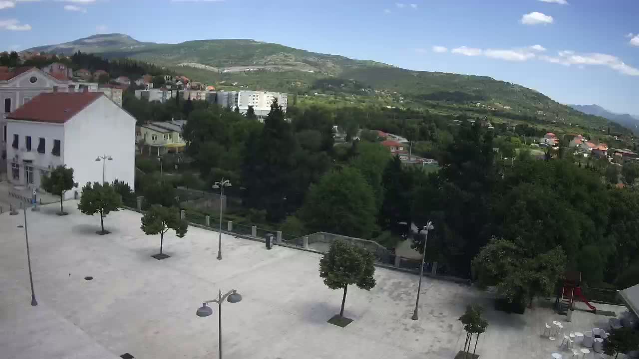 A paved square with sparsely planted trees overlooks a town nestled in a valley beneath a partly cloudy sky and mountains in the background.