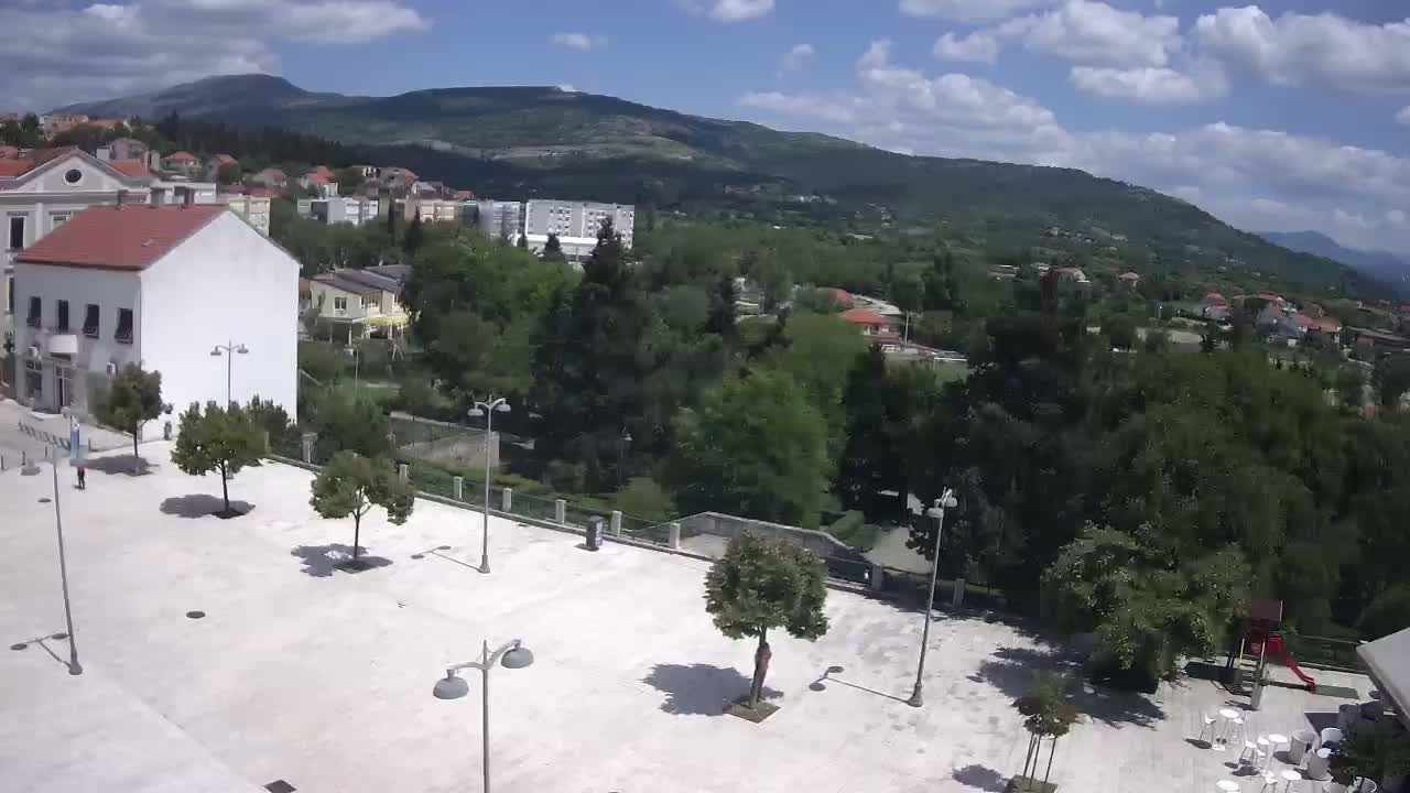 A mostly sunny day reveals a town square with several trees and lampposts, overlooking a verdant hillside and distant mountains.