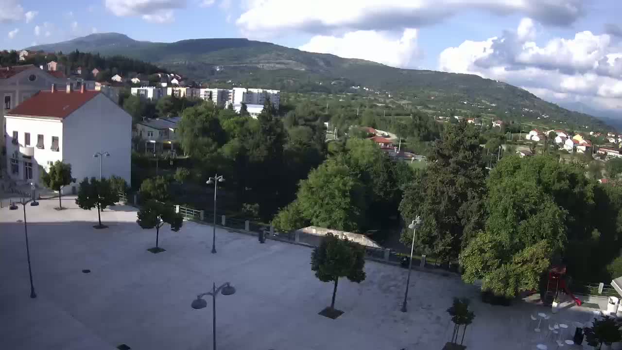 A mostly sunny day reveals a town square with several trees and buildings, overlooking a verdant valley and distant mountains.