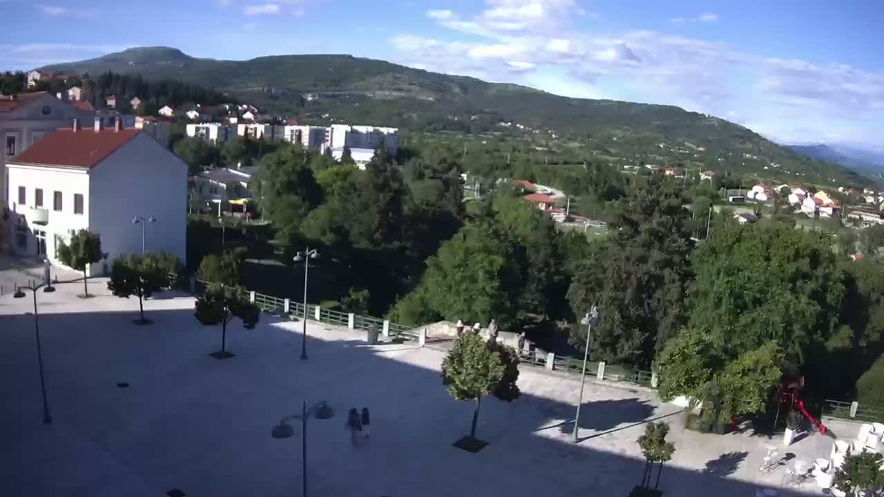 A mostly sunny day overlooking a town square with several buildings and trees, situated at the base of a mountain range.