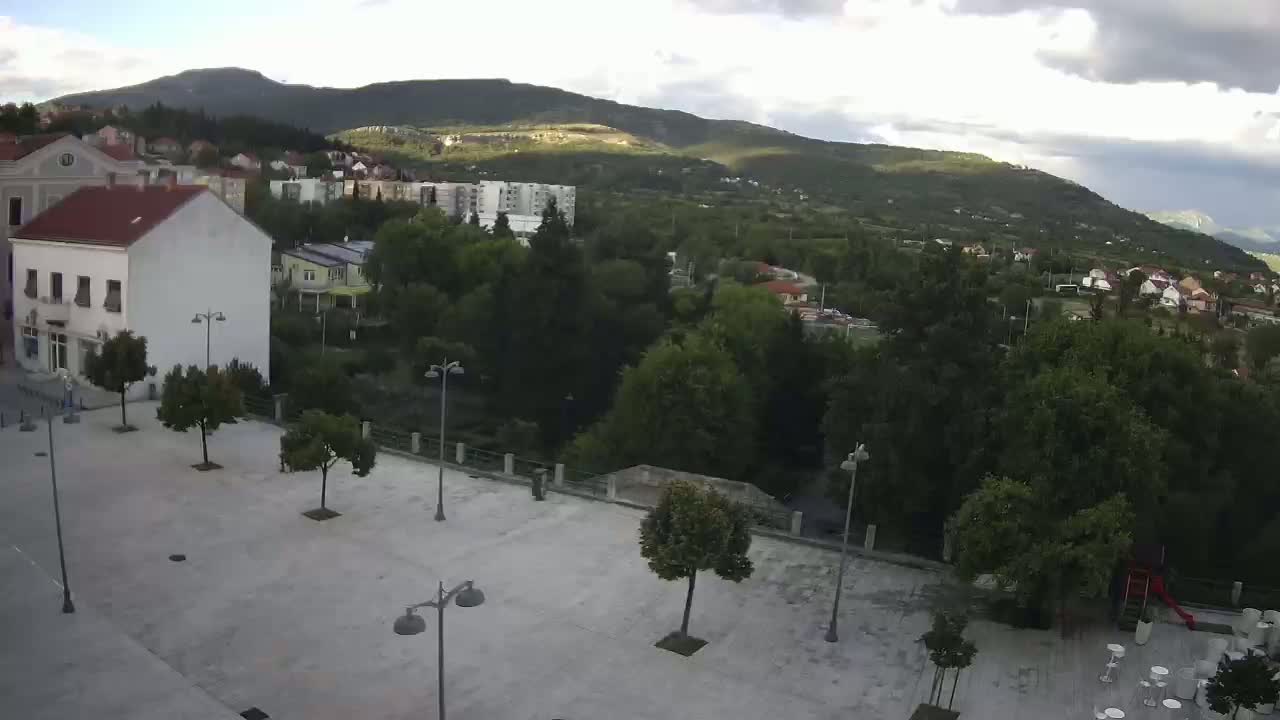 A mostly empty town square with a few trees and lampposts, overlooks a lush green valley and mountains under partly cloudy skies.