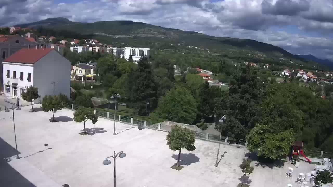 An empty town square with a few trees and some outdoor furniture is situated below a hillside town under a partly cloudy sky.