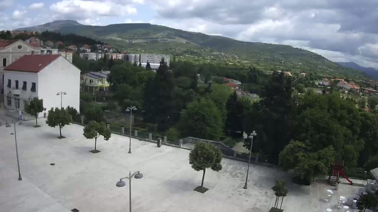 An empty town square with several trees and streetlights is situated below a hill with lush green vegetation under a partly cloudy sky.