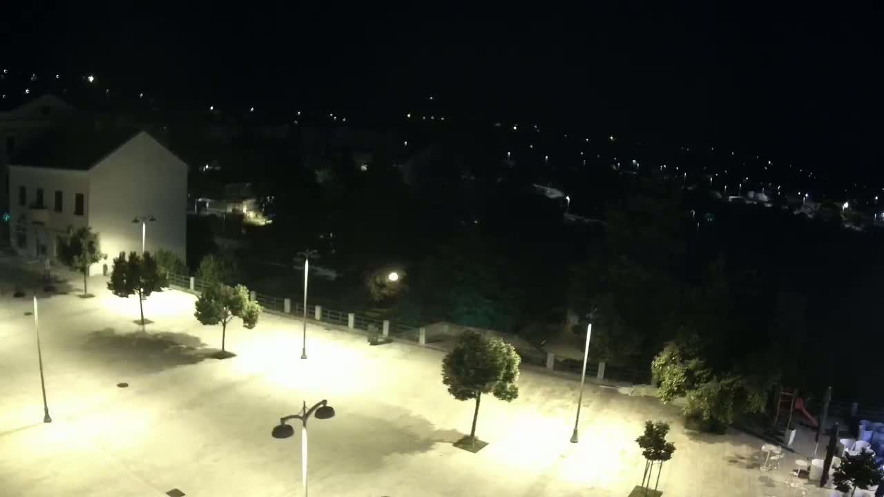 A brightly lit, empty town square at night, surrounded by trees and buildings, overlooks a darker, tree-lined area with distant lights.