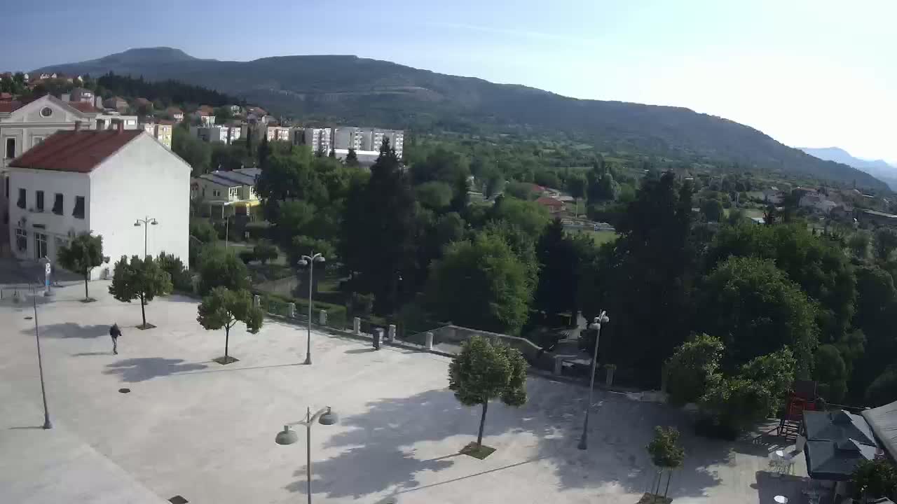 A mostly empty town square with a few pedestrians and trees, situated in front of buildings and backed by a verdant hillside under a clear sky.