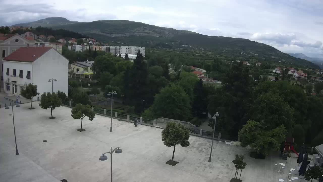 A mostly empty paved town square with several small trees and lampposts, overlooks a hillside town nestled beneath a mountain range on an overcast day.