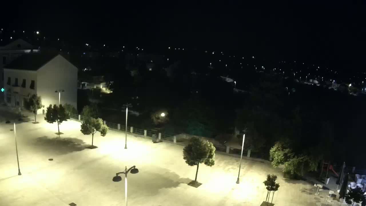 A brightly lit plaza at night, surrounded by trees and buildings, overlooks a darker, sparsely lit town under a clear sky.
