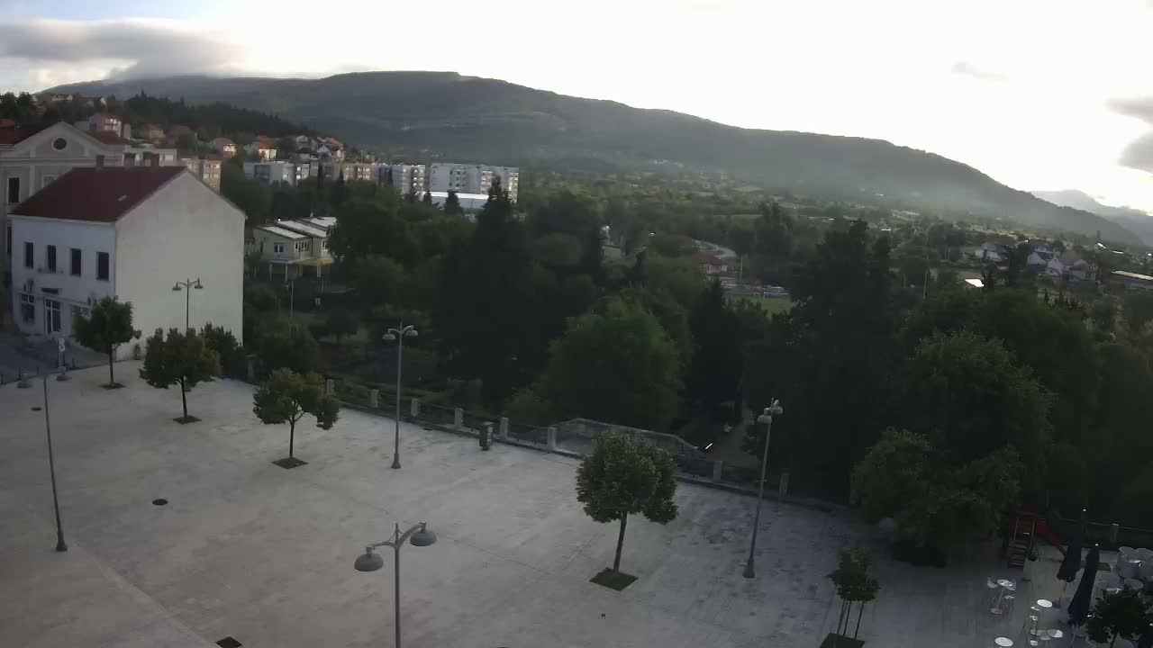 A mostly empty paved square with several young trees and lampposts is situated in front of buildings, overlooking a town nestled in a valley beneath hazy mountains under a partly cloudy sky.