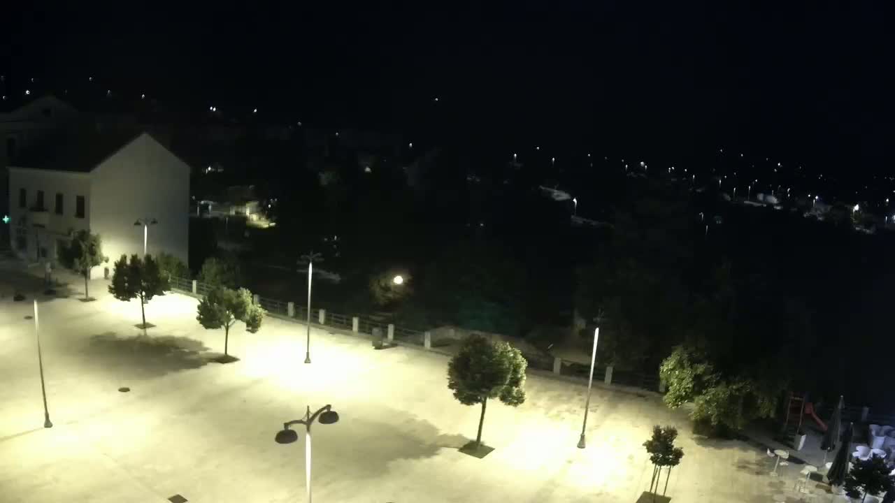 A nighttime, high-angle view of a brightly lit plaza with several trees and lampposts, adjacent to a building and a dark body of water with faint lights in the distance.