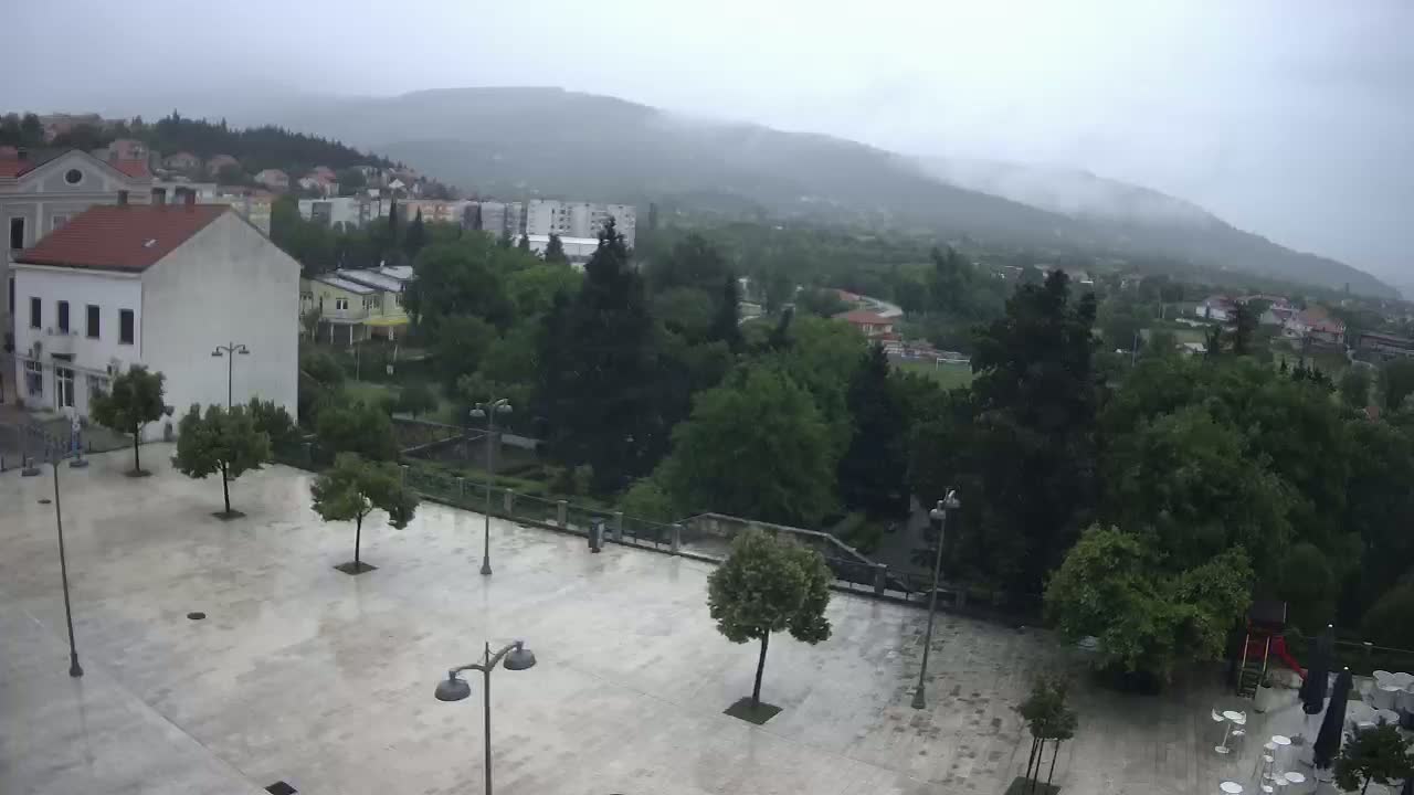 A wet, paved town square with several trees and buildings, overlooks a hilly landscape shrouded in mist.