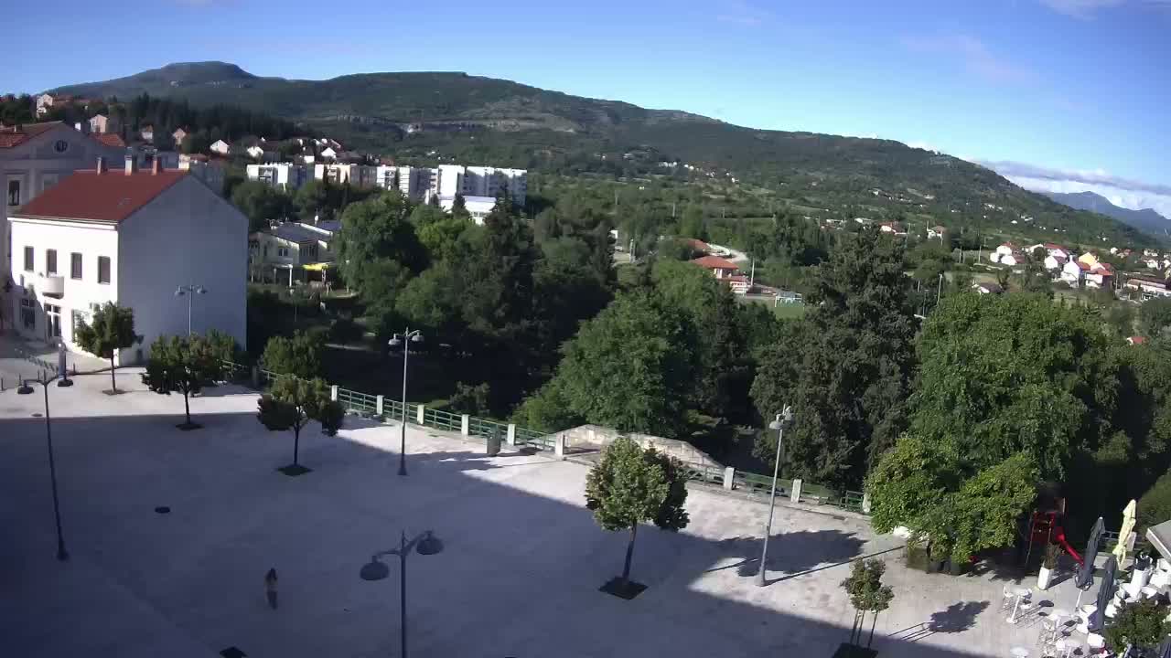 A mostly sunny day reveals a town square overlooking a bridge and a verdant valley, with a mountain range in the background.