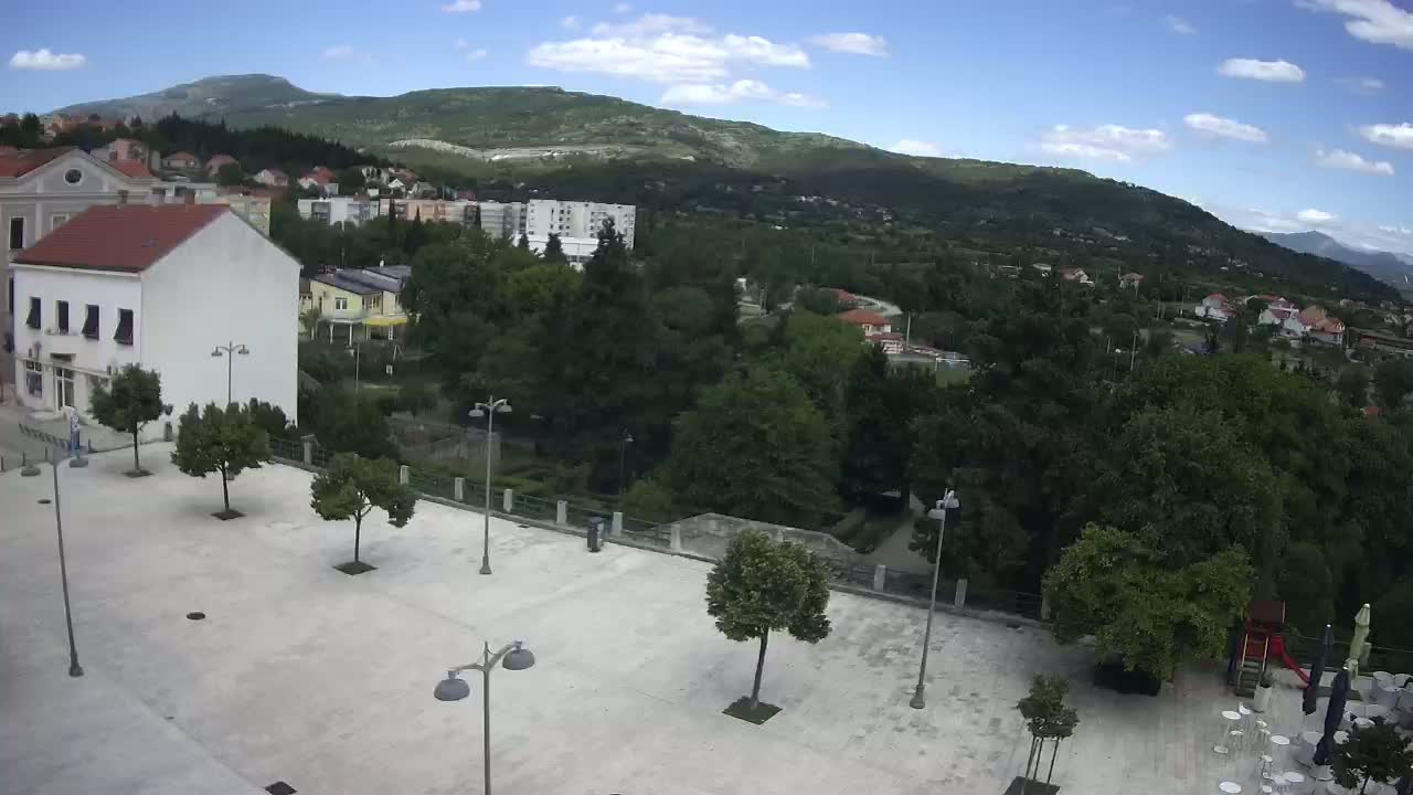 A paved town square with sparsely placed trees and lampposts, overlooking a verdant hillside under a mostly sunny sky.
