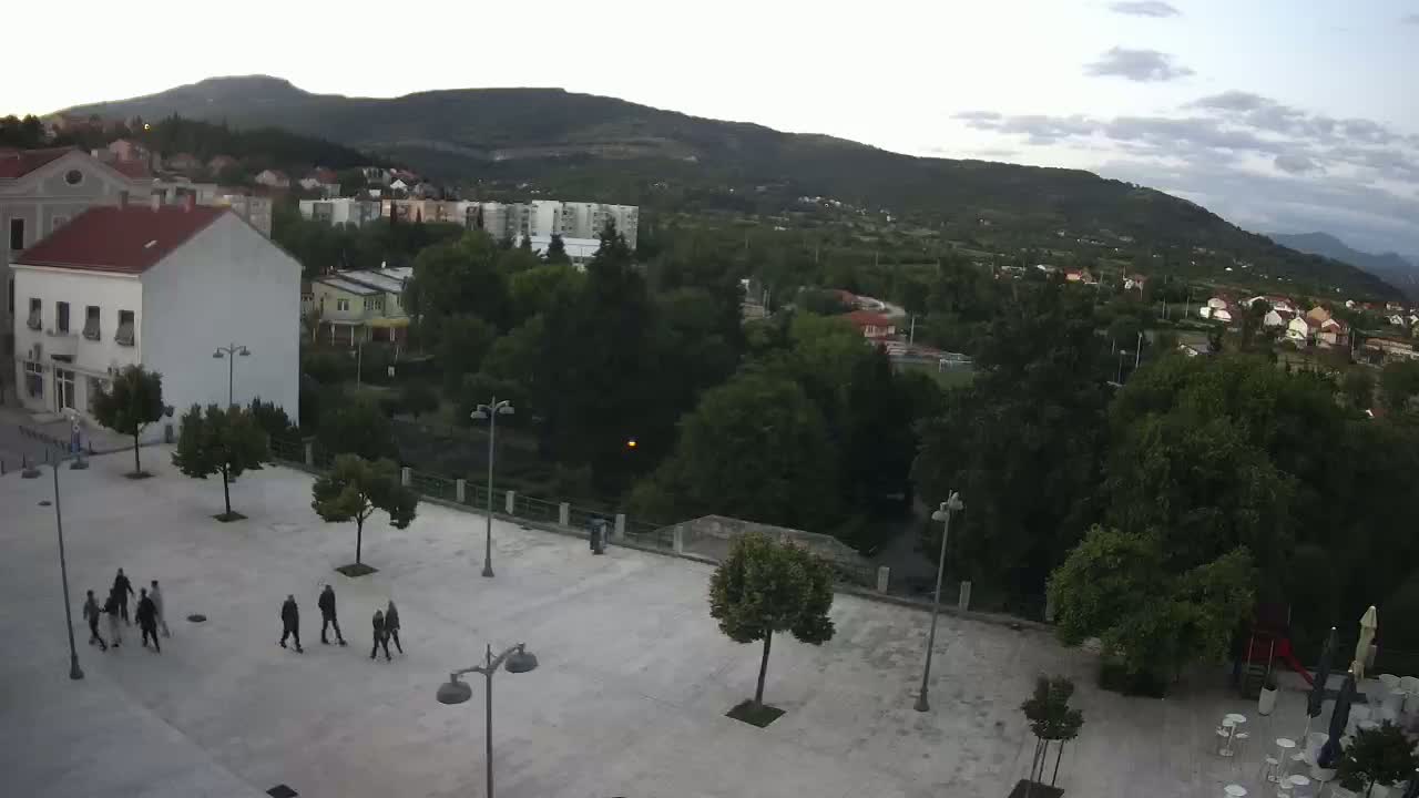 An elevated view of a town square with several people walking, surrounded by buildings and lush green trees under a partly cloudy sky.