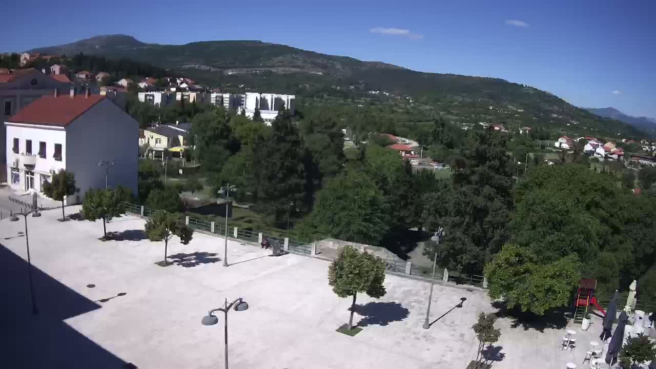 A sunny day reveals a town square with several trees, a few buildings, and a playground, all situated below a mountain range.