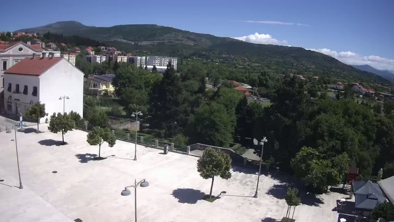 An empty, light-colored plaza with several trees and lampposts is situated in front of buildings, overlooking a town nestled amongst green hills under a clear, sunny sky.
