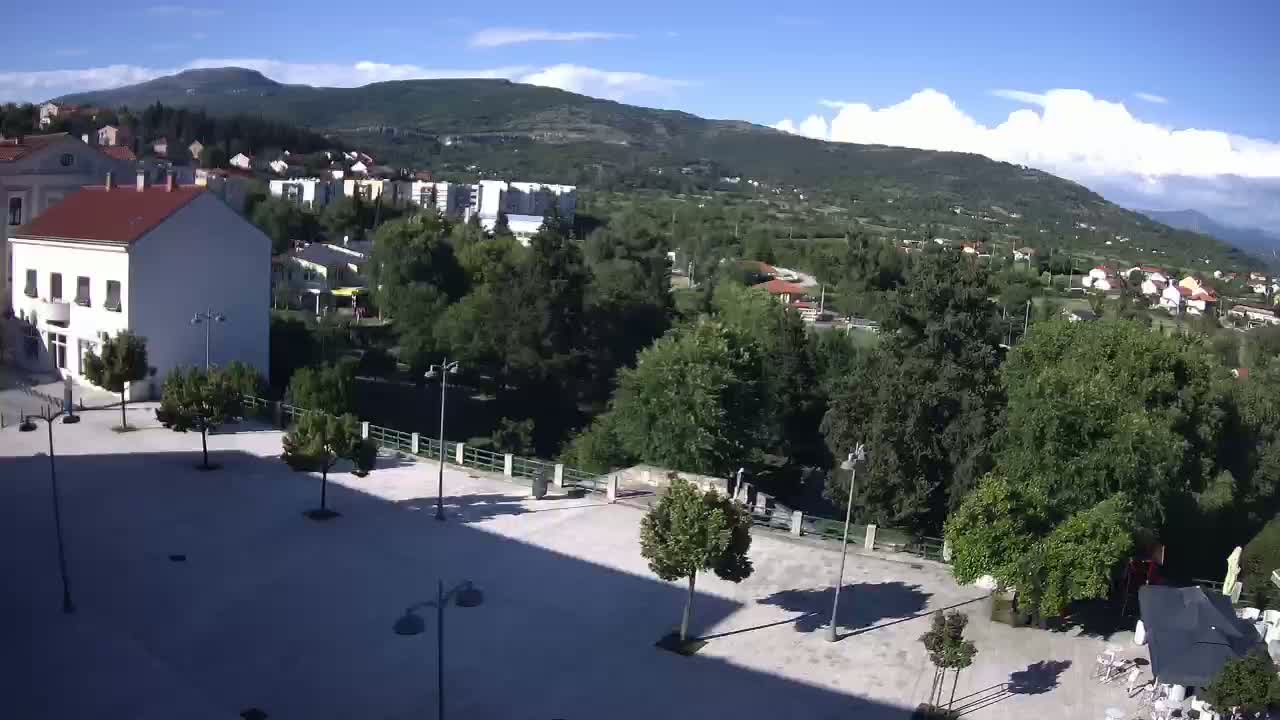A mostly sunny day reveals a town square, buildings, and lush green hills under a partly cloudy sky.