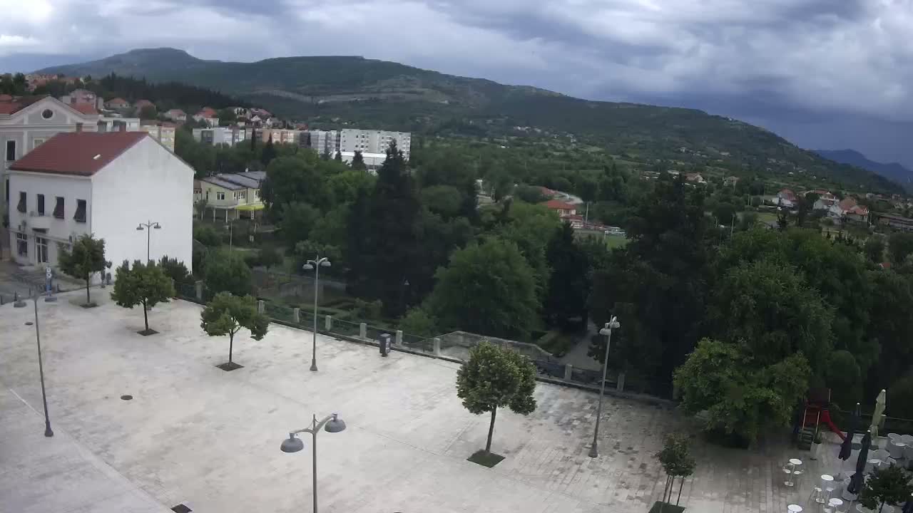 An empty town square with lampposts and a few trees under an overcast sky overlooks a hilly green landscape.