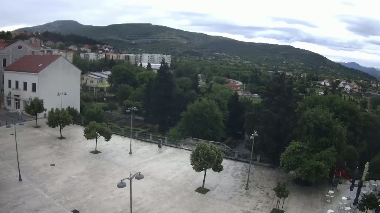 An empty town square with several small trees and lampposts is overlooked by buildings and lush green hills under a cloudy sky.