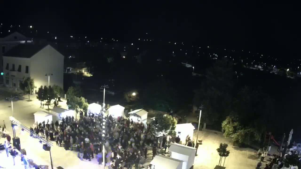 A large crowd gathers in a town square at night under clear skies, surrounded by small kiosks and buildings.