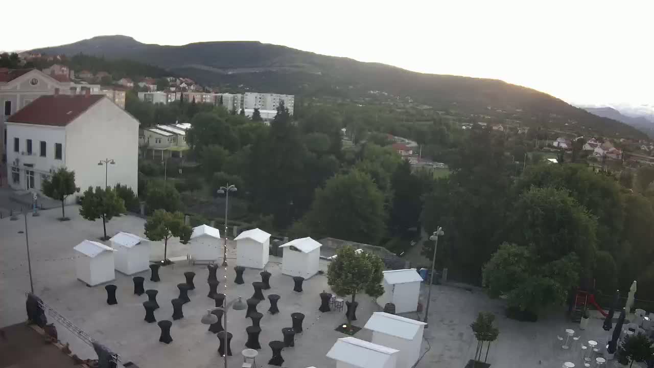 An aerial view of a town square with small white structures and numerous black tables set up, surrounded by lush green trees and buildings under a clear, bright sky.